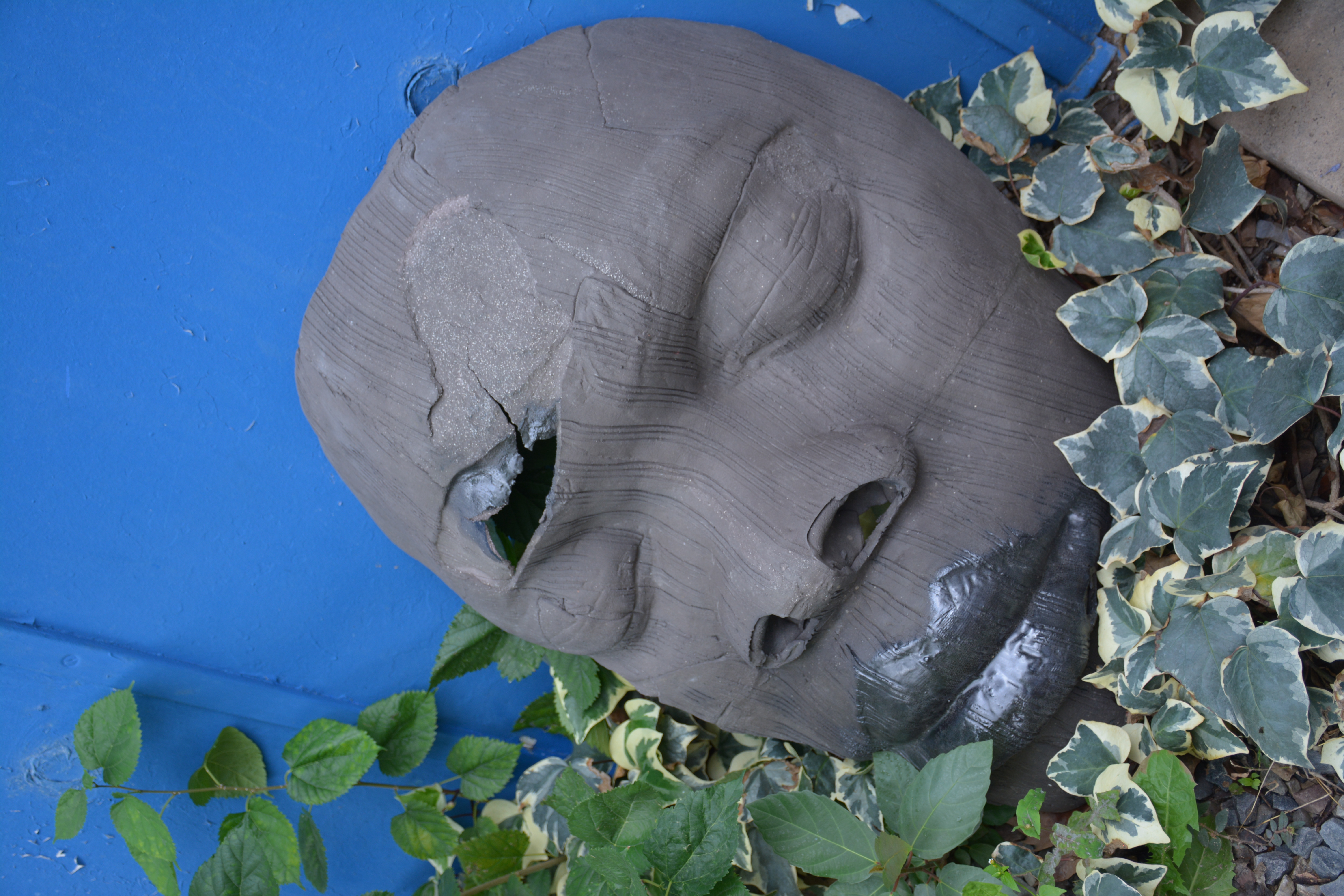 Damaged sculptural face resting among leaves against a blue wall